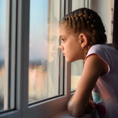 A child with her hair tied back is leaning against and looking out of the window