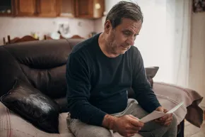Photograph of a man sat on a sofa looking worried whilst reading a letter